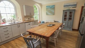 A kitchen with a wooden table and chairs, white cabinets, and a window with a view of greenery. - Beggars Roost (Truro)
