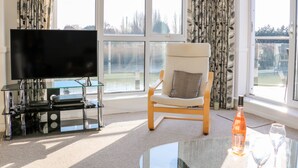 Een moderne woonkamer met een flatscreen televisie, een glazen salontafel, een comfortabele fauteuil en uitzicht op een park door grote ramen.