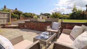 An outdoor patio with wicker furniture, a stone wall, and a view of a garden. - Coney Garth Cottage (Beckermet)