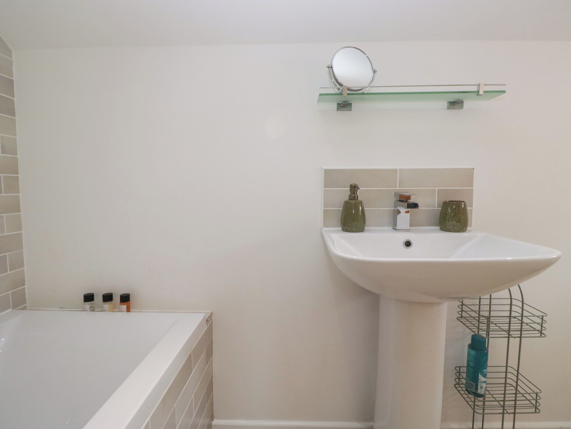 Ein Badezimmer mit einer weißen Badewanne, einem weißen Waschbecken mit grünem Seifenspender und einem Spiegel auf einer Glasablage.