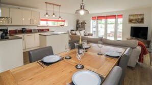 A modern kitchen with white cabinets, a dining area with a wooden table set for four, and a living area with a sofa and a television. - Coney Garth Cottage (Beckermet)