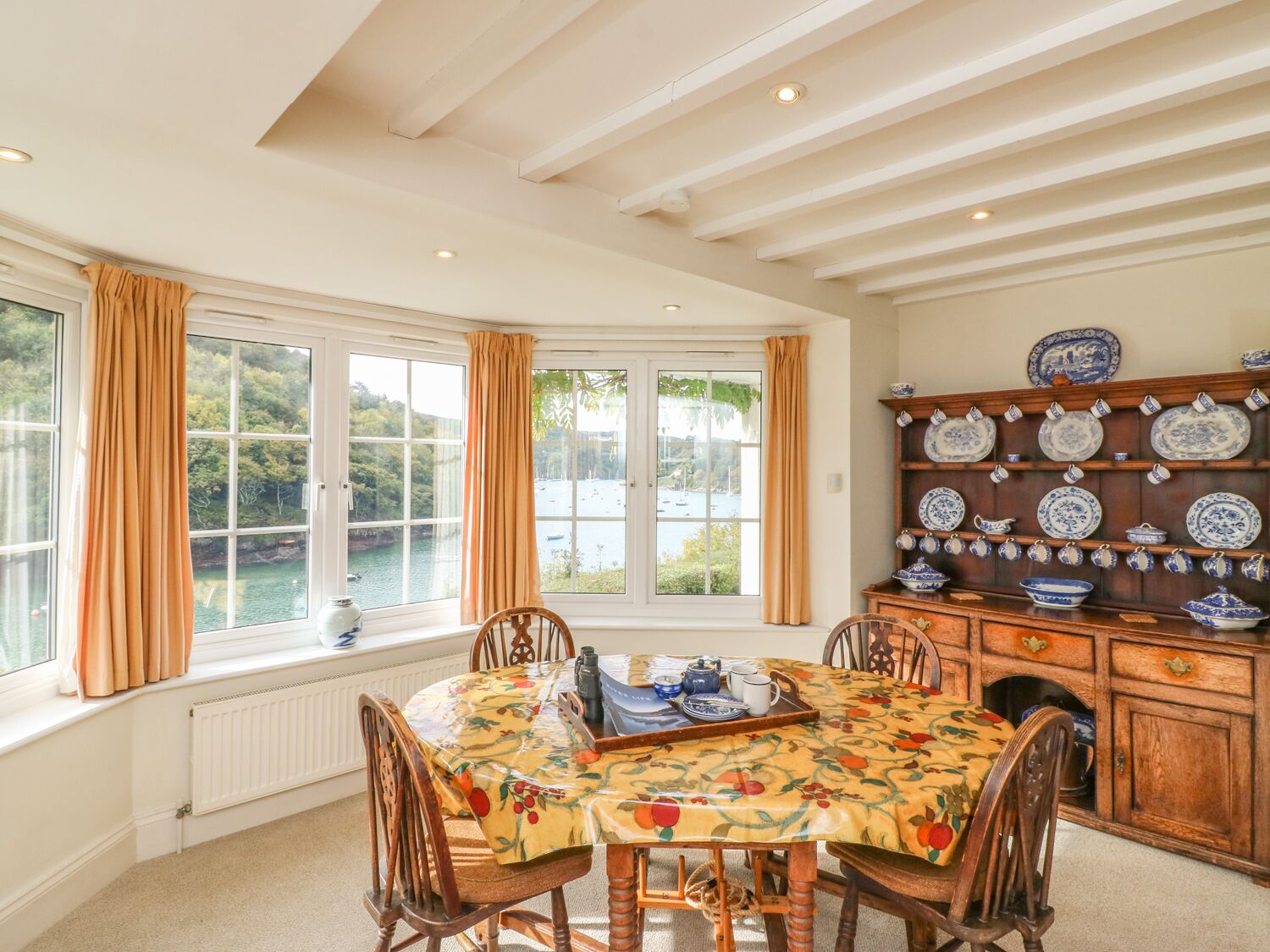 Un coin repas avec une table dressée pour quatre personnes, entourée de chaises en bois. On y trouve une grande fenêtre avec des rideaux, un radiateur en dessous, et un buffet en bois contenant de la vaisselle bleue et blanche.