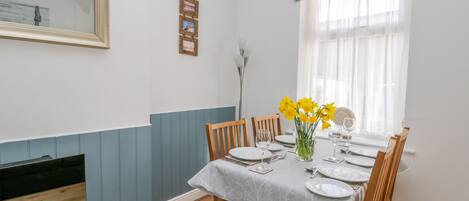 A dining area with a table set for four, wooden chairs, a vase with yellow flowers, and a window with a blue-striped valance.