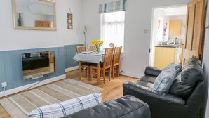A living room with a black leather sofa, a dining table with chairs, a TV mounted on the wall, and a kitchen area visible through an open door. - Sea Breakers Cottage (Filey)