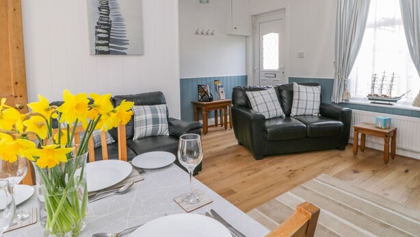 A cozy living room with a black leather sofa, a dining table set with plates and a vase of yellow flowers, and a window with curtains. - Sea Breakers Cottage (Filey)