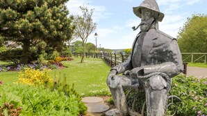 A statue of a seated figure in a park with a fence, greenery, and a pathway. - Sea Breakers Cottage (Filey)