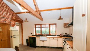 A kitchen with a brick wall, wooden beams, a window, and a sink. - Honey Buzzard Barn (Fakenham)