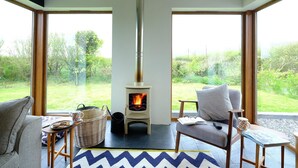 Une pièce chaleureuse avec un poêle à bois, de grandes fenêtres, des sièges confortables et une vue sur l’extérieur.