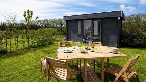 Une petite cabane en bois sombre, avec une table et des chaises placées à l’extérieur.