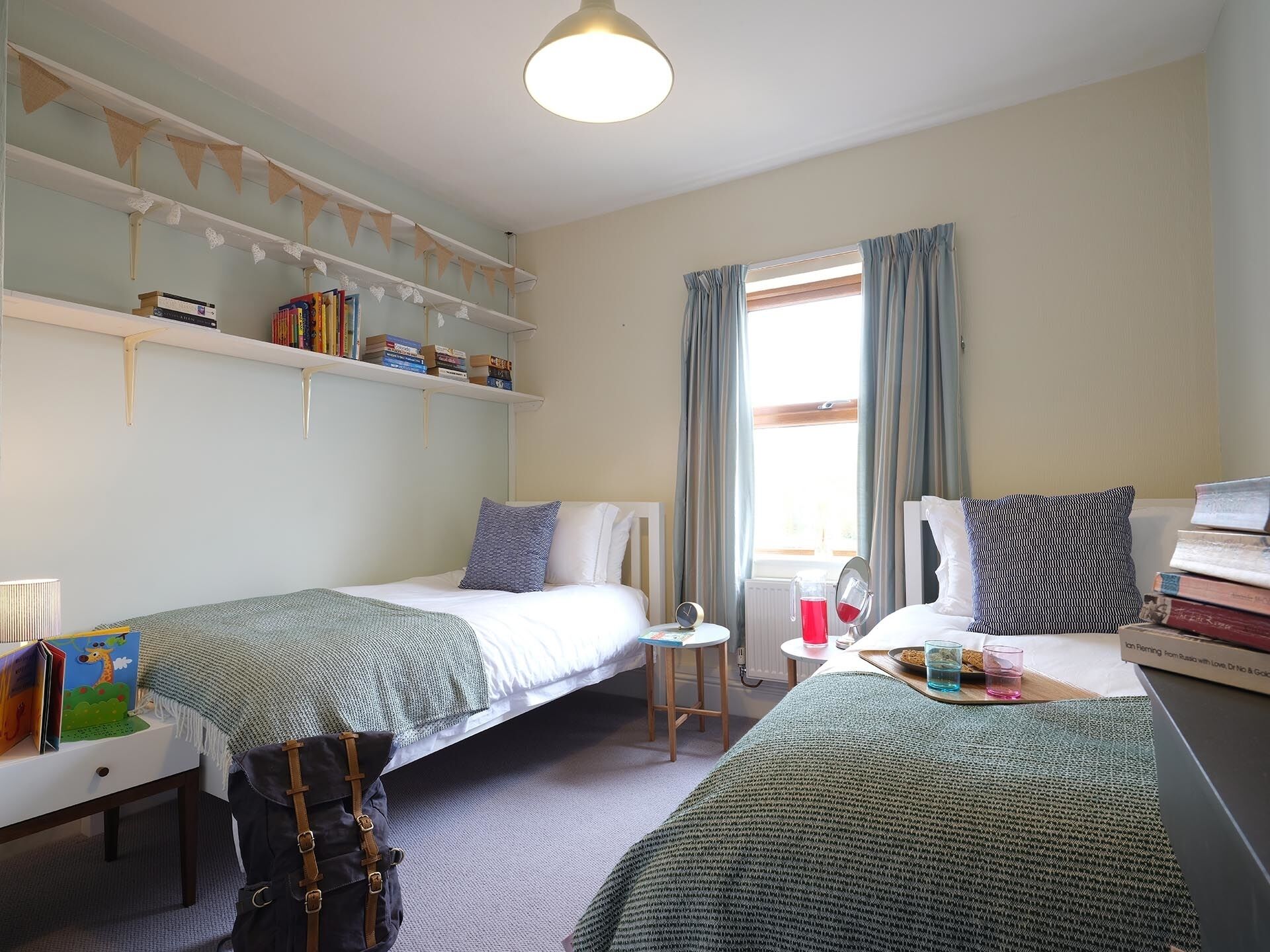 Un dormitorio acogedor con dos camas individuales, una mesita con una silla, una estantería y una ventana con cortinas.