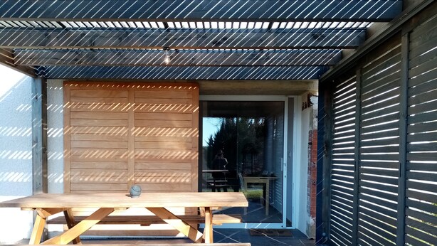 A wooden picnic table with a bench, a metal roof, and a garage door in the background. - La Gurlanne (Lasseran)