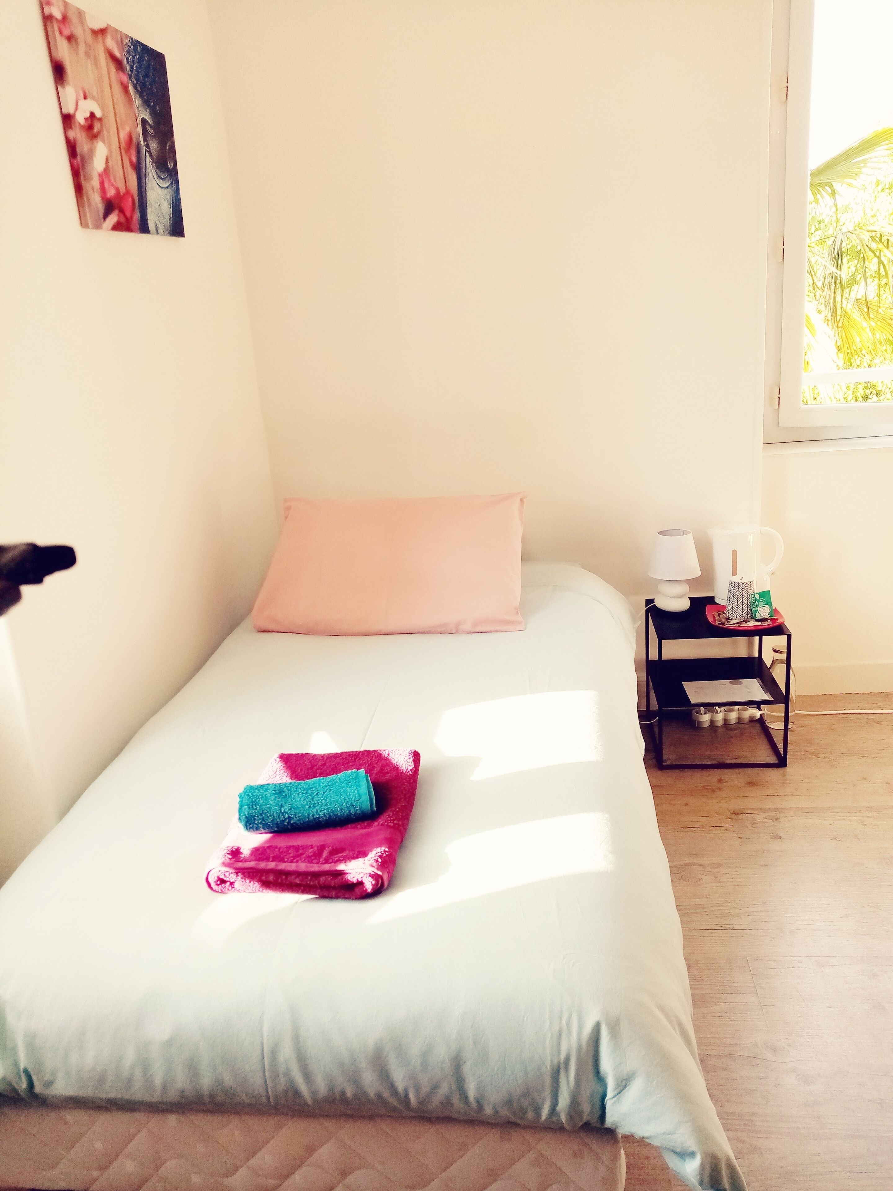 A neatly made bed with a light blue bedspread, a pink pillow, and folded towels. There is a small black nightstand with a lamp and a mug beside the bed.
