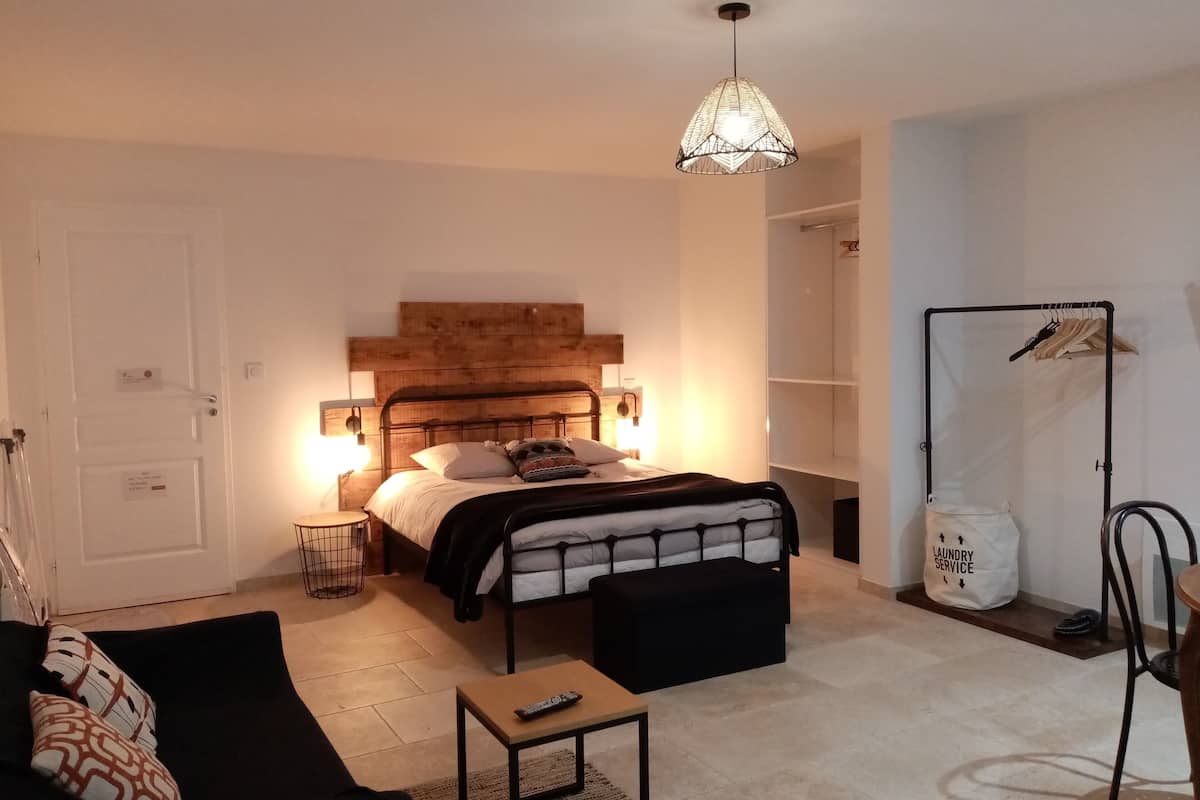 A bedroom with a wooden headboard, a bed with a black and white patterned bedspread, a black bench, a small table with a remote control, a hanging light fixture, and a laundry service bag.