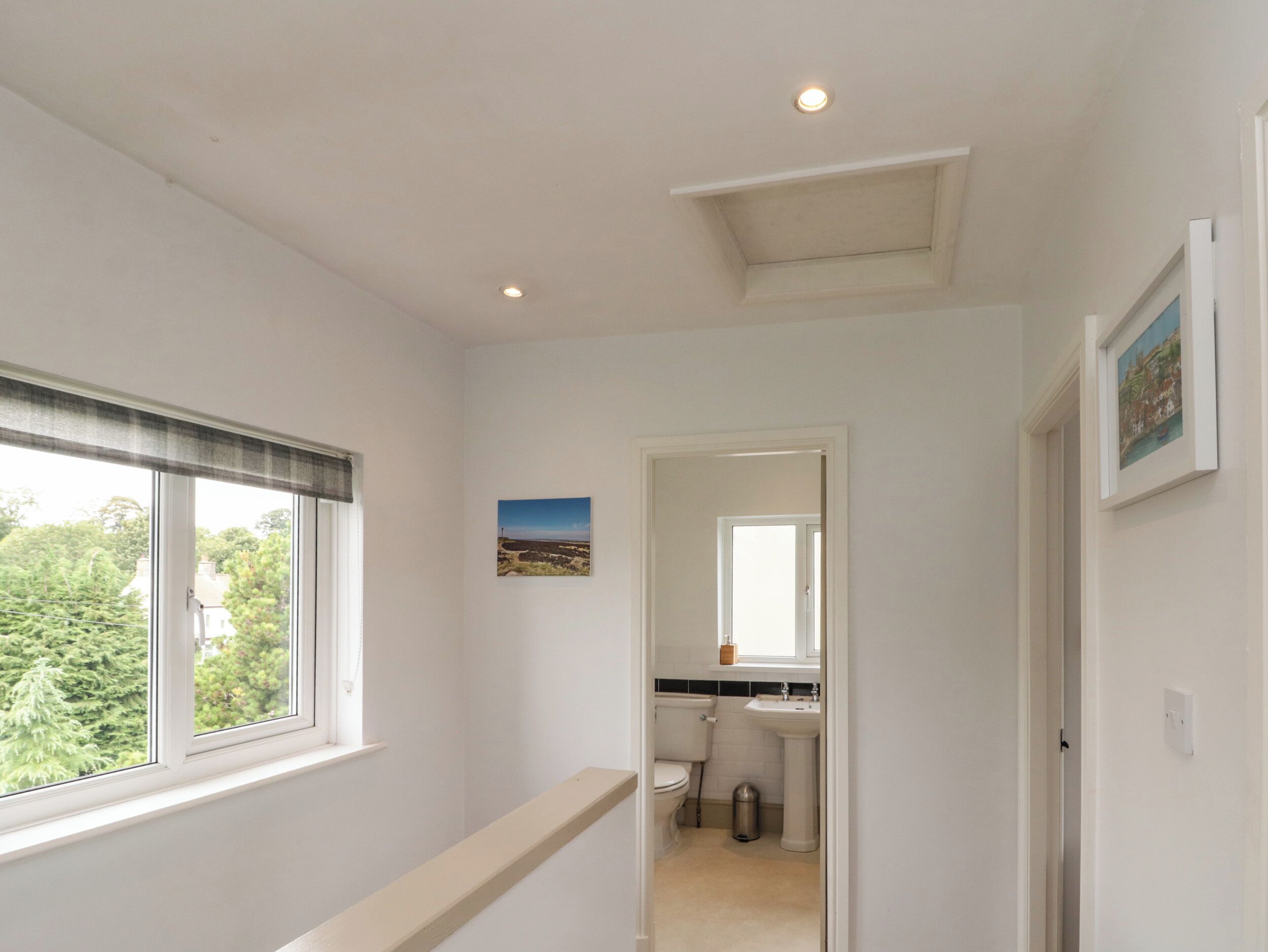 A hallway with a window, a picture on the wall, and a view of a bathroom.
