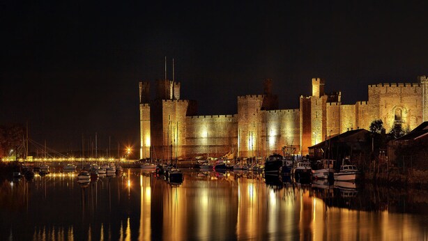 A historic castle with a moat, illuminated towers, and boats docked in the water.