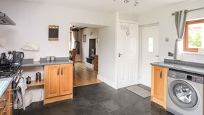 A compact kitchen with a stove, kettle, and washing machine, wooden cabinets, and a tiled floor.