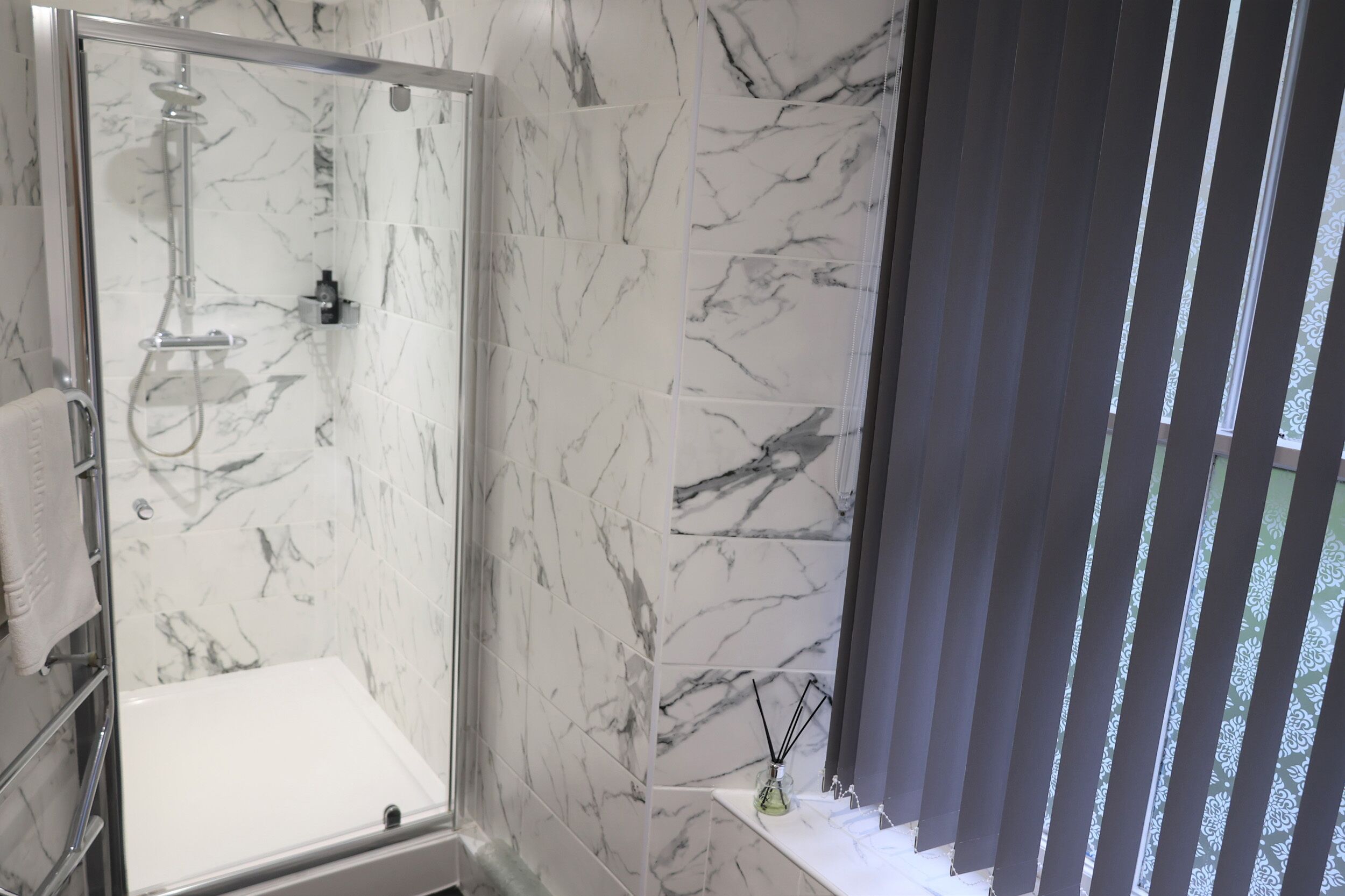 Un baño con ducha envuelta en vidrio, baldosas de mármol, un lavabo con espejo y una ventana con persianas.
