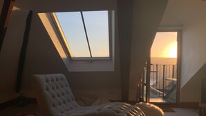 A room with a sloped ceiling, a large window, a cushioned chair, a small table, and a balcony with a railing.