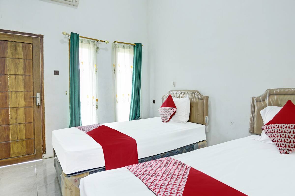 Una habitación de hotel con dos camas, cada una con una colcha roja y blanca, una puerta de madera y una ventana con cortinas verdes.