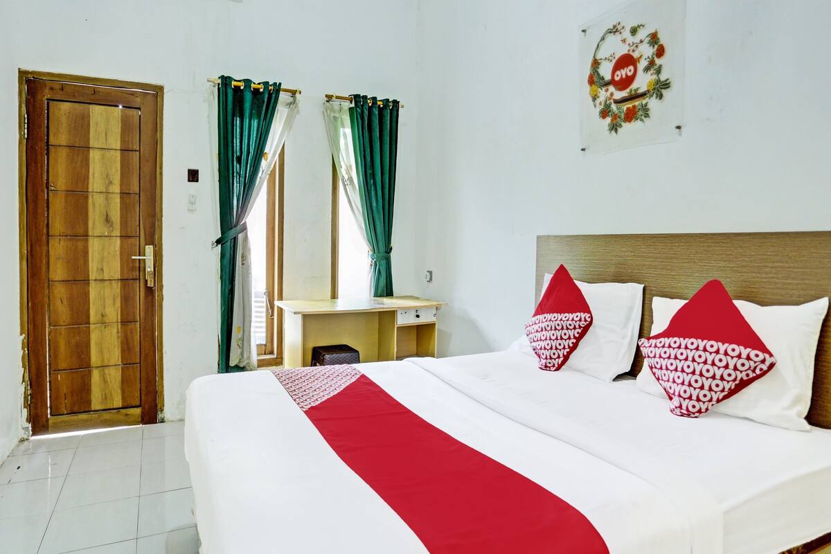 Una habitación de hotel con puerta de madera, un escritorio con silla, una cama con ropa de cama blanca y roja, y una decoración en la pared.