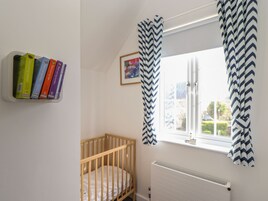 Una habitación con una cuna, un radiador, una ventana con cortinas en zigzag y una estantería de pared con libros de colores.