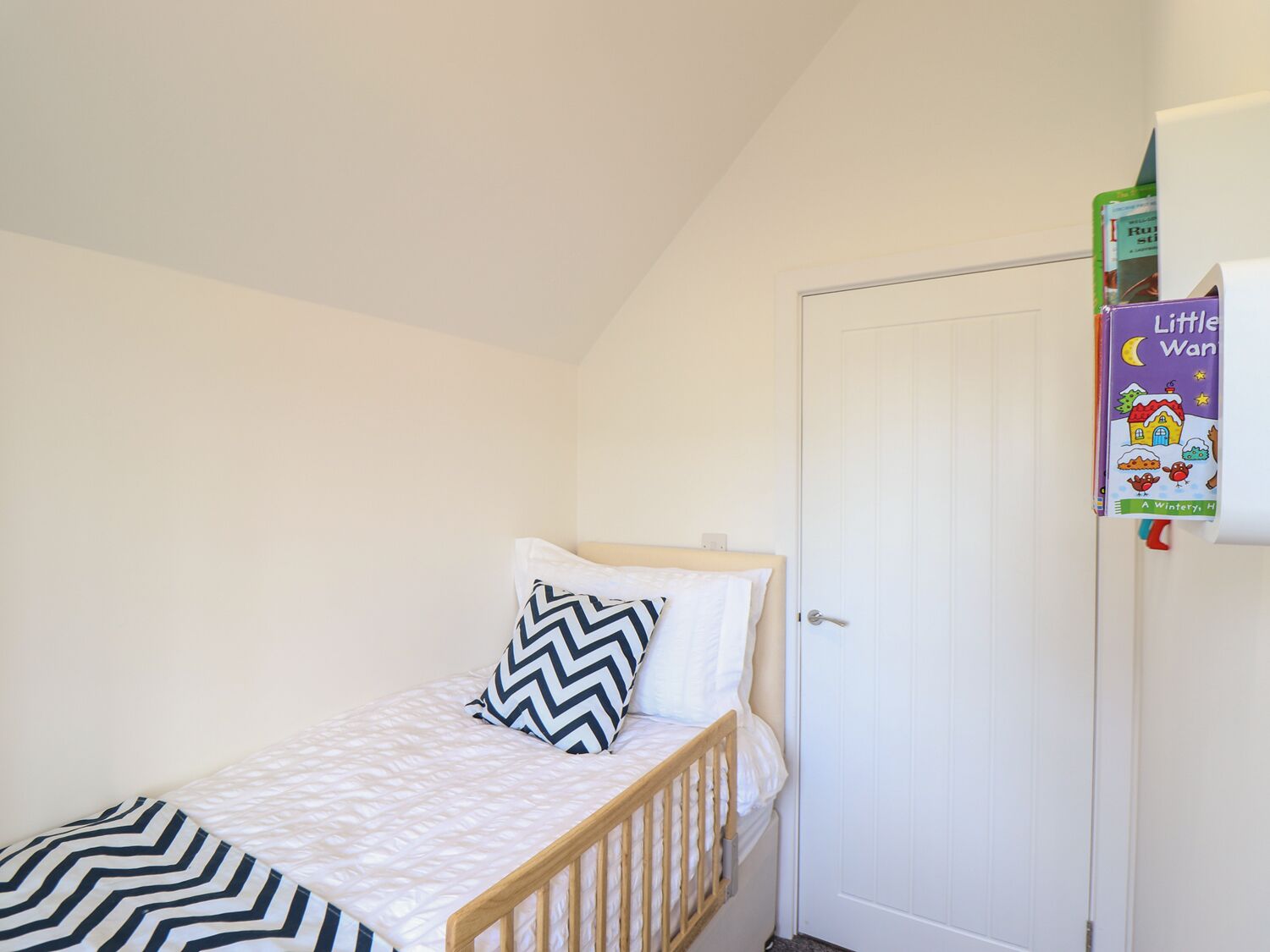 Une petite chambre avec un lit simple, un cadre en bois, un matelas blanc et un oreiller à motifs chevrons. On aperçoit une porte blanche, et un livre pour enfants est accroché au mur.