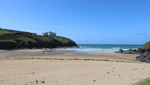 Un paisaje costero con una playa arenosa, un edificio blanco grande sobre una colina cubierta de césped y el océano al fondo.