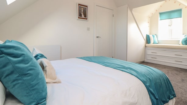 A bedroom with a sloped ceiling, a bed with teal bedding, a white dresser, and a window with a view. - Kober House (Helston)