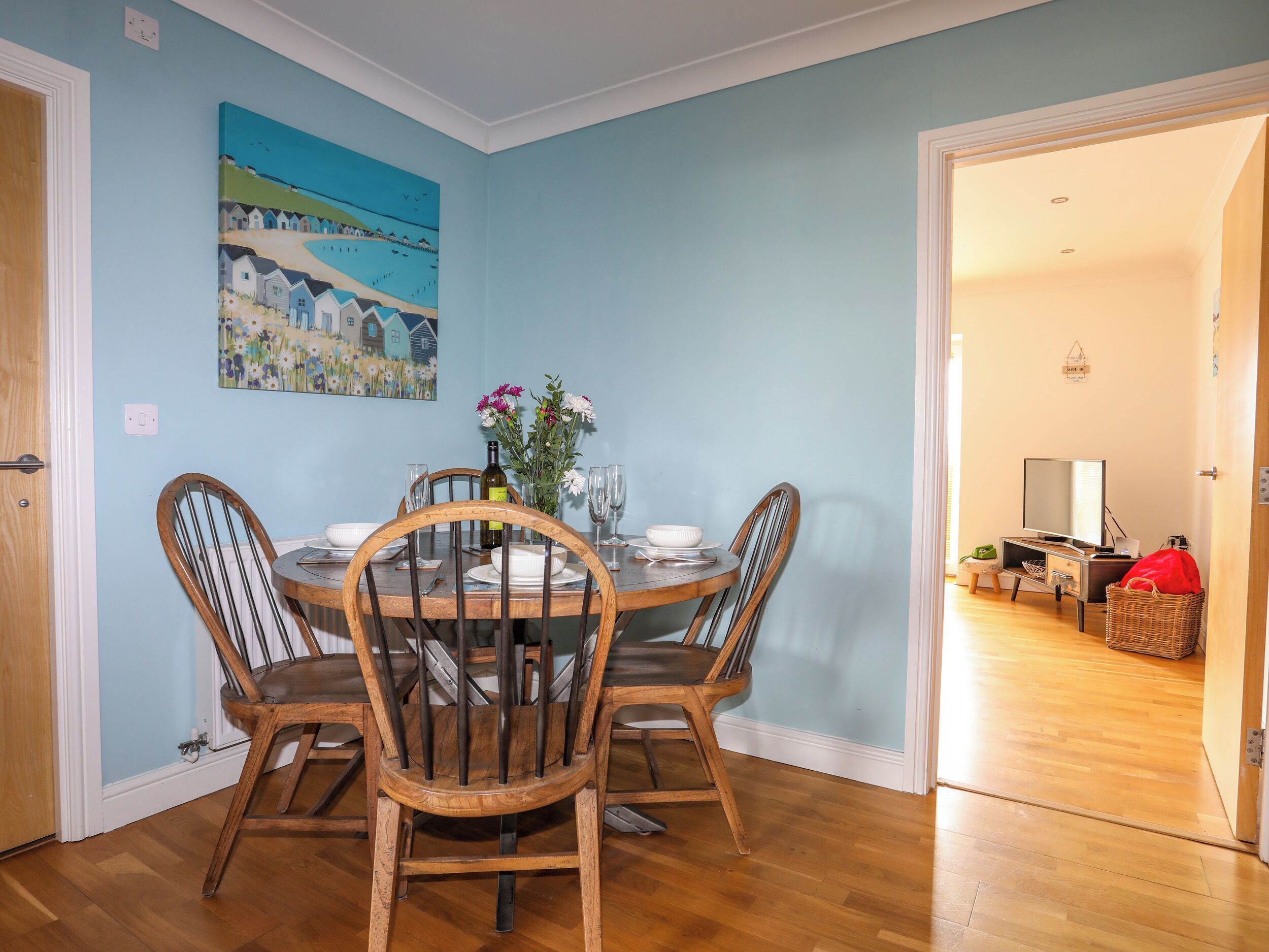 A dining area with a round table, four wooden chairs, a vase with flowers, and a painting on the wall.