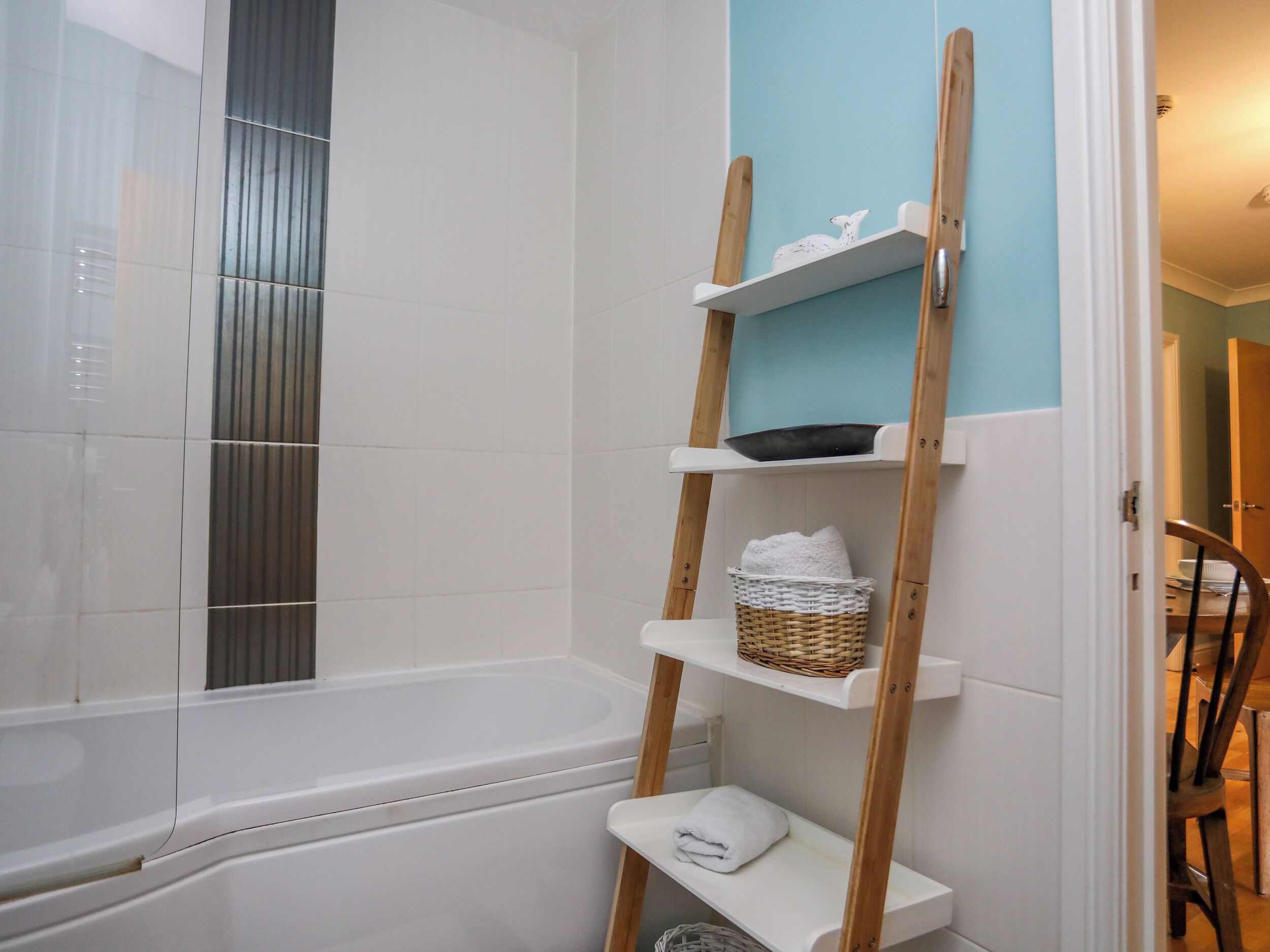 Une salle de bain avec une baignoire blanche, une étagère en bois avec un panier et une serviette, et un mur bleu.