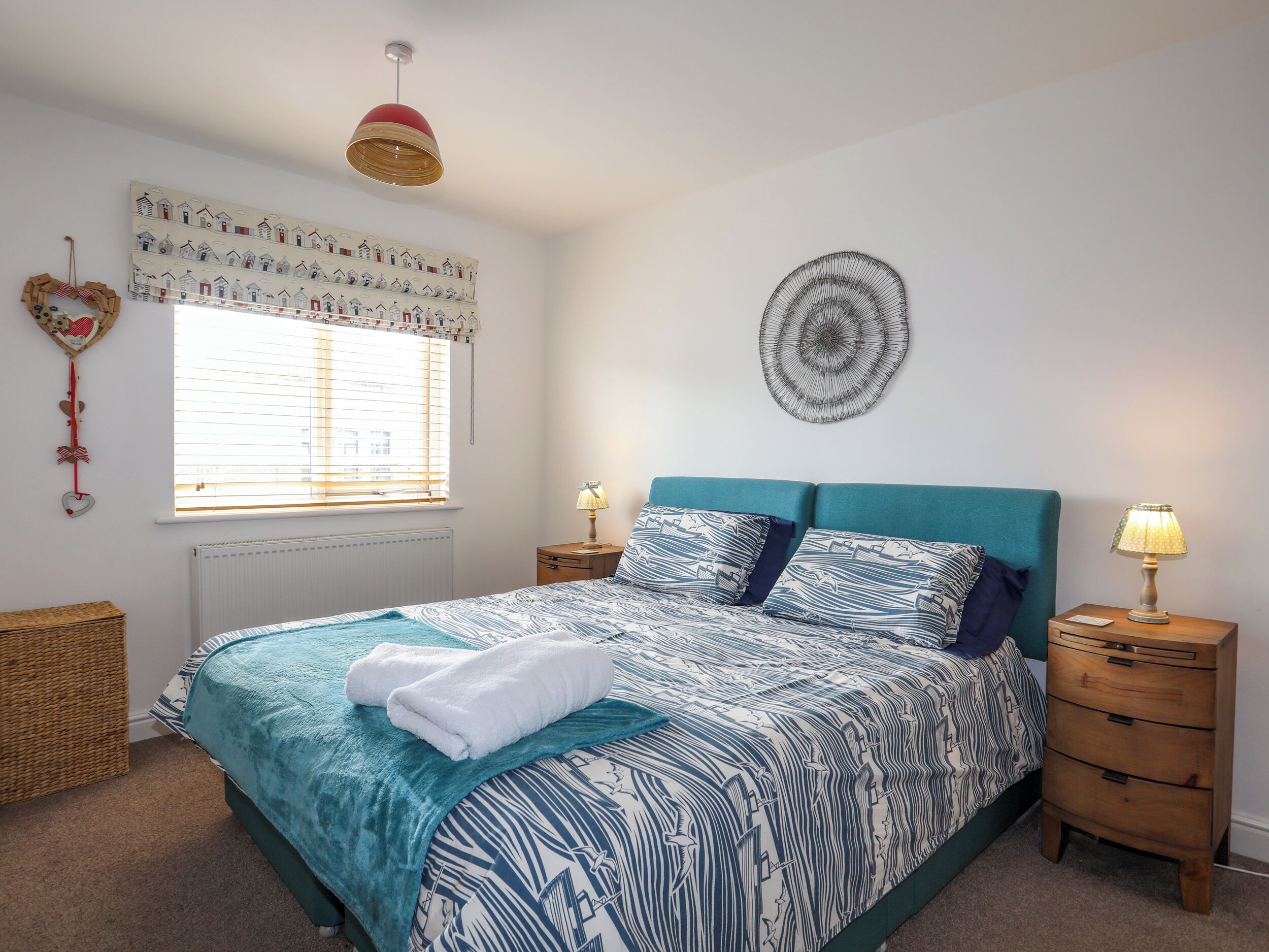 A bedroom with a bed, two bedside tables, a window with blinds, a wall hanging, and a ceiling light.