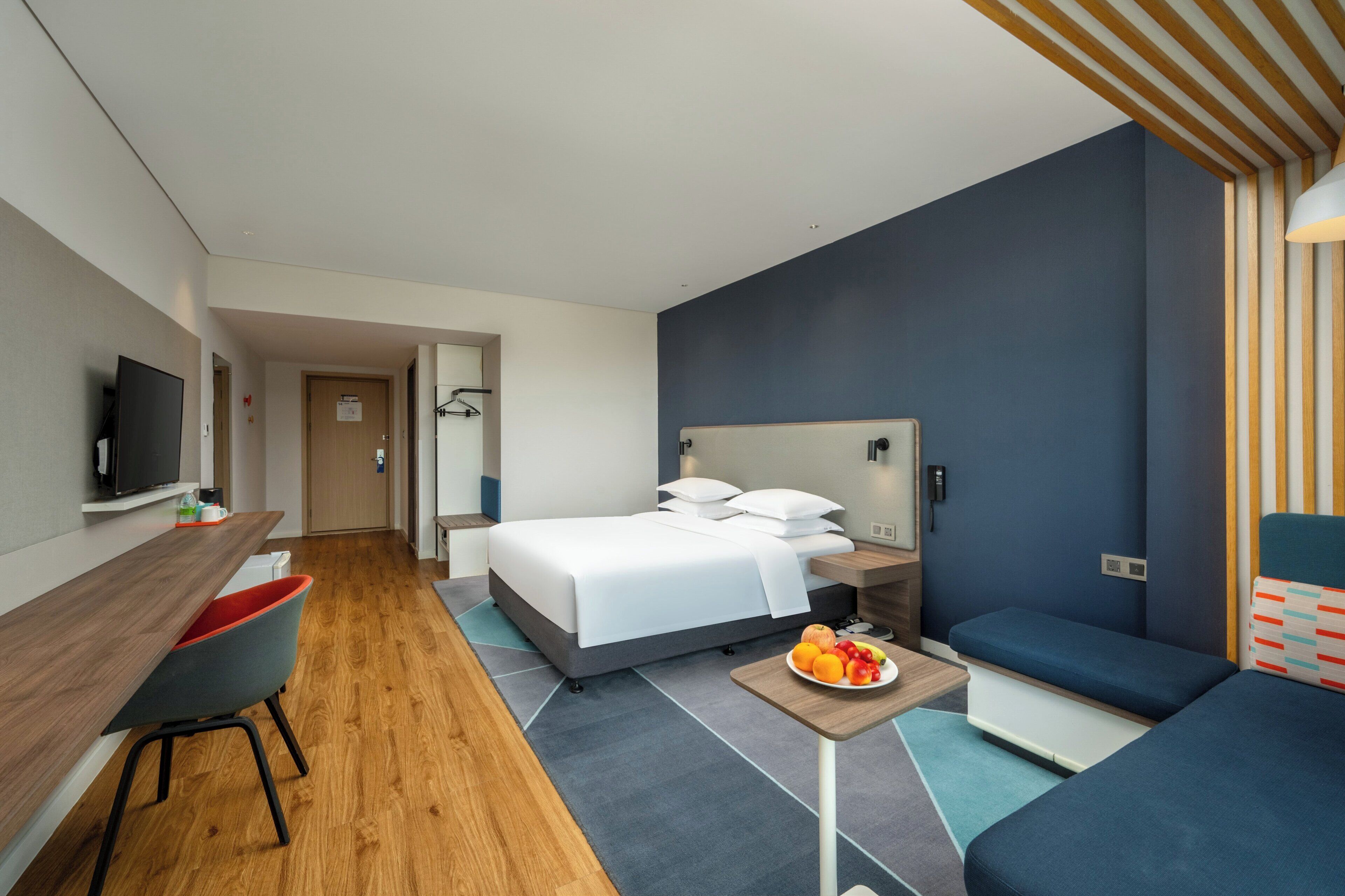 Una habitación de hotel moderna con cama, mesita de noche, televisor y una zona de estar con sofá y sillón.