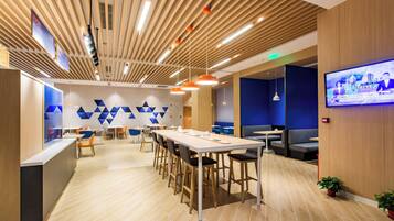 A modern interior with a long communal table, individual seating, and a television screen.