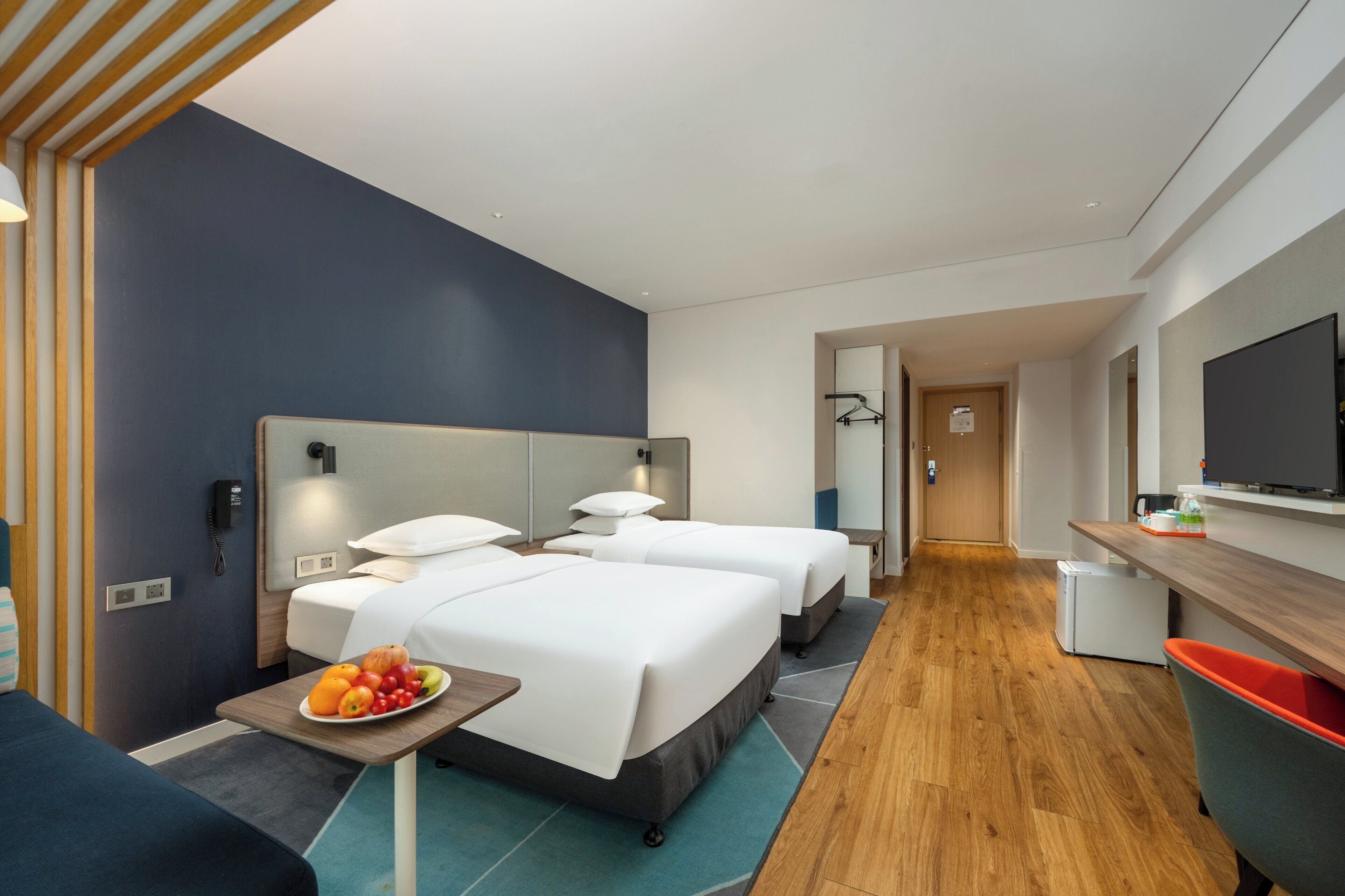 A modern hotel room with two beds, a desk, a TV, and a fruit bowl on the side table.