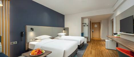 A modern hotel room with two beds, a desk, a TV, and a fruit bowl on the side table.