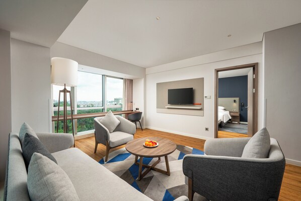 A modern living room with a sofa, armchairs, a coffee table with fruit, a TV mounted on the wall, and a large window offering a city view. - Holiday Inn Express Jiangmen Jianghai by IHG (Jiangmen)