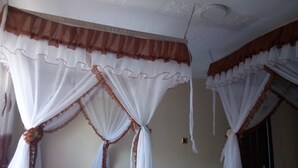 A room with a bed, white curtains with brown trim, and a ceiling with faintly visible text. - Hungry & T Hippo (Naivasha)