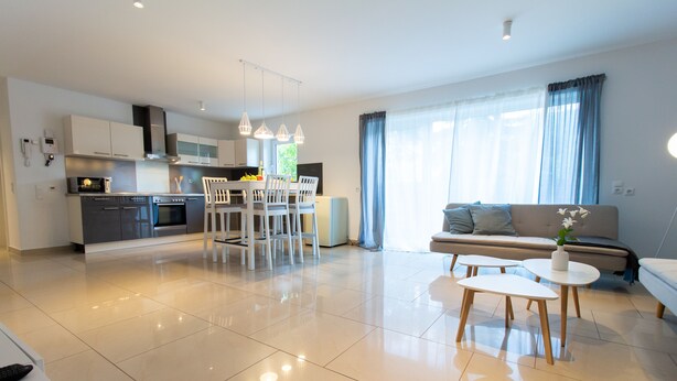 A modern kitchen with white cabinets, a dining area with a white table and chairs, and a living area with a sofa and a small coffee table. - T & T Suites Raunheim (Raunheim)