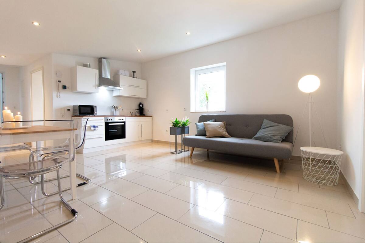 A modern kitchen with white cabinetry, a dining area with a glass table and chairs, a grey sofa, and a floor lamp.