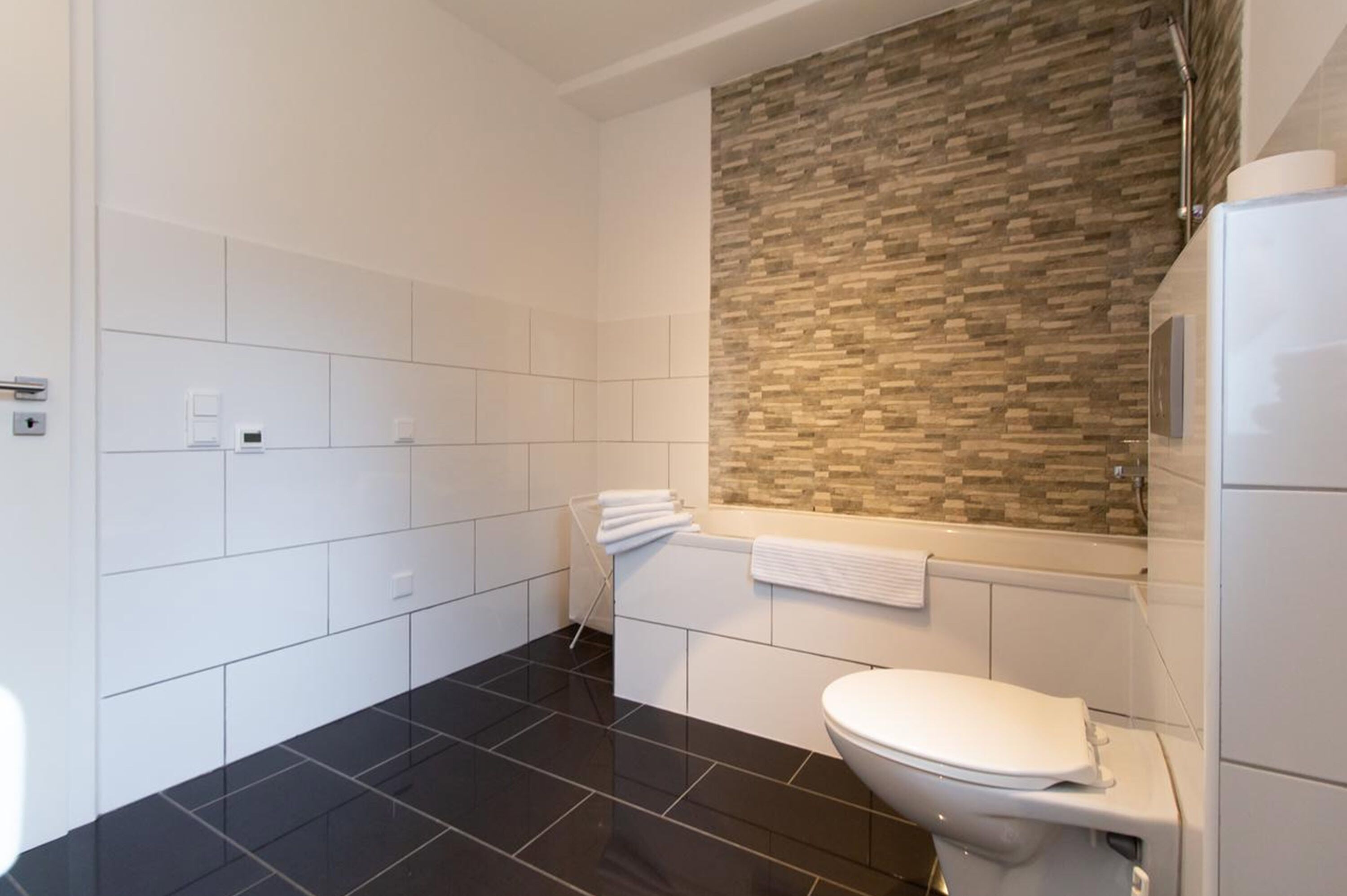A modern bathroom with a black tiled floor, a white toilet, and a wall with a textured stone backsplash.
