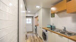 Una cocina compacta con paredes de azulejos blancos, muebles de madera, una lavadora y un lavamanos.