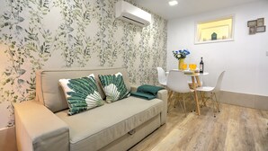 Una sala de estar moderna con un sofá color beige, almohadones blancos y verdes estampados, una mesita con un jarrón de flores y un aire acondicionado instalado en la pared.