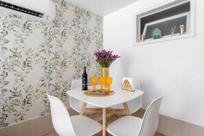 A dining area with a white table, four white chairs, a vase with purple flowers, a bottle of wine, and two golden goblets.