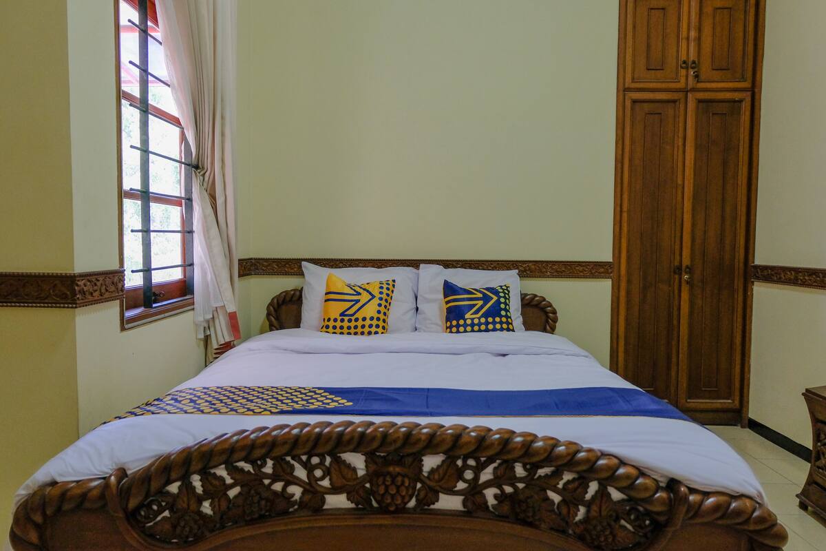 Quarto com estrutura de cama de madeira, duas almofadas com padrões geométricos, janela com cortinas e guarda-roupa de madeira.