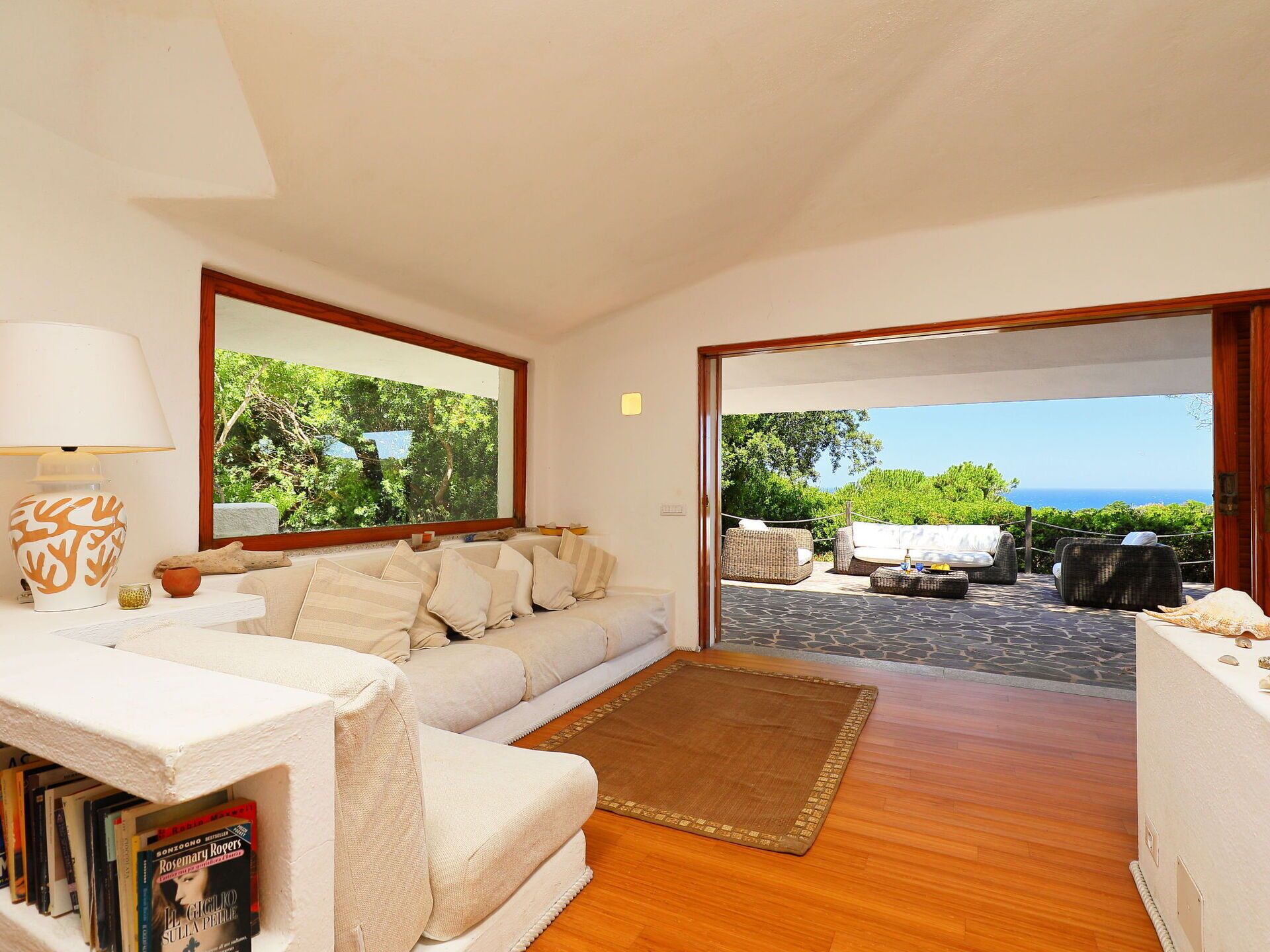 Ein gemütliches Wohnzimmer mit einer Couch, einem Couchtisch und einem Teppich. Ein Bücherregal mit Büchern und eine Lampe sind ebenfalls vorhanden. Große Fenster bieten Blick auf Grünflächen und das Meer.