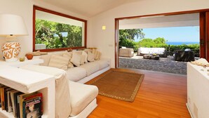 A cozy living room with a sofa, a coffee table, and a rug. There is a bookshelf with books and a lamp. The room has large windows with a view of greenery and the sea. - Beautiful private villa for 7 people with WIFI, TV, terrace and pets allowed (Aglientu)