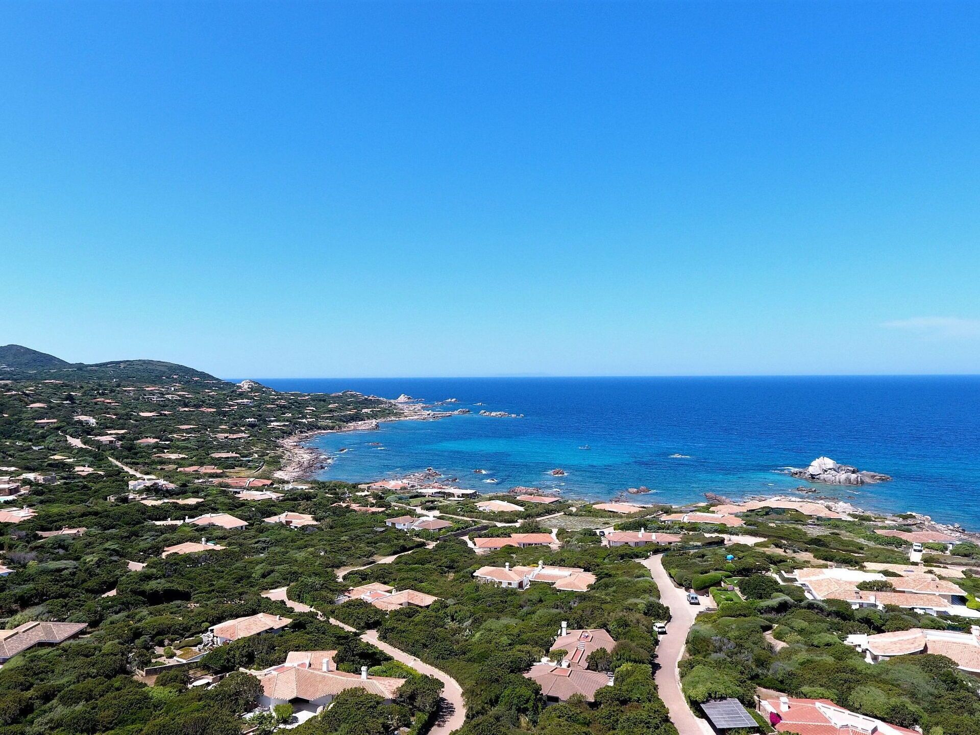 An aerial view of a coastal residential area with houses, roads, and a clear blue sea.