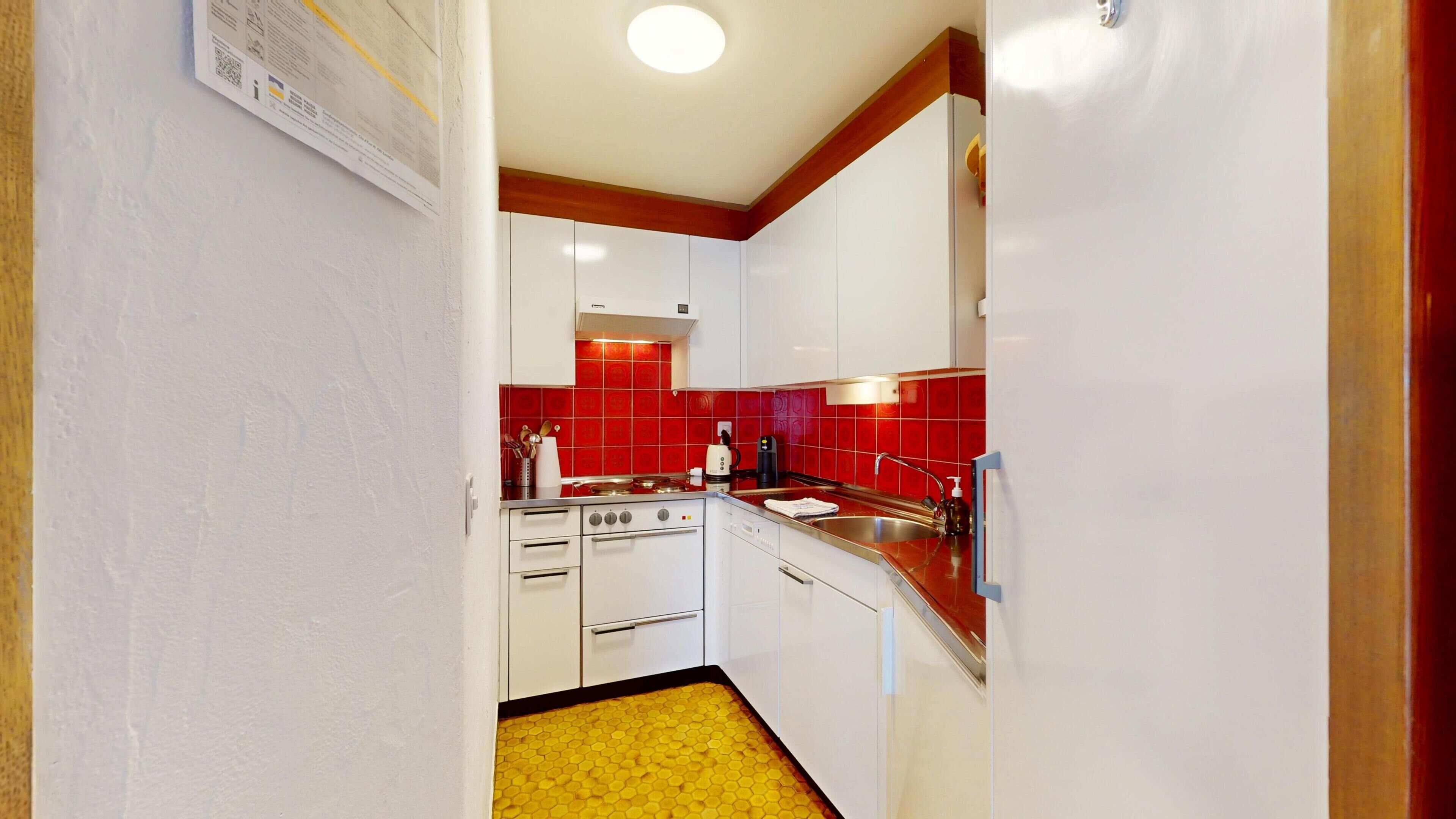 A compact kitchen with white cabinets, red tiles, a yellow hexagonal floor, and a white stove.