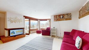 Un salón con televisión, sofá rojo, alfombra estampada y vistas a montañas a través de ventanas amplias.