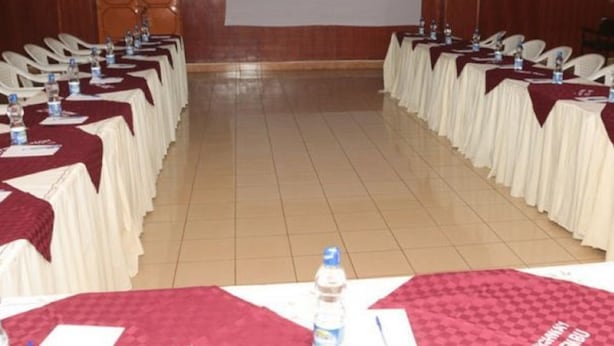 A conference room with rows of tables, each with a white tablecloth and a red table runner. Water bottles and papers are on the tables. - Maina Highway Hotel (Embu)